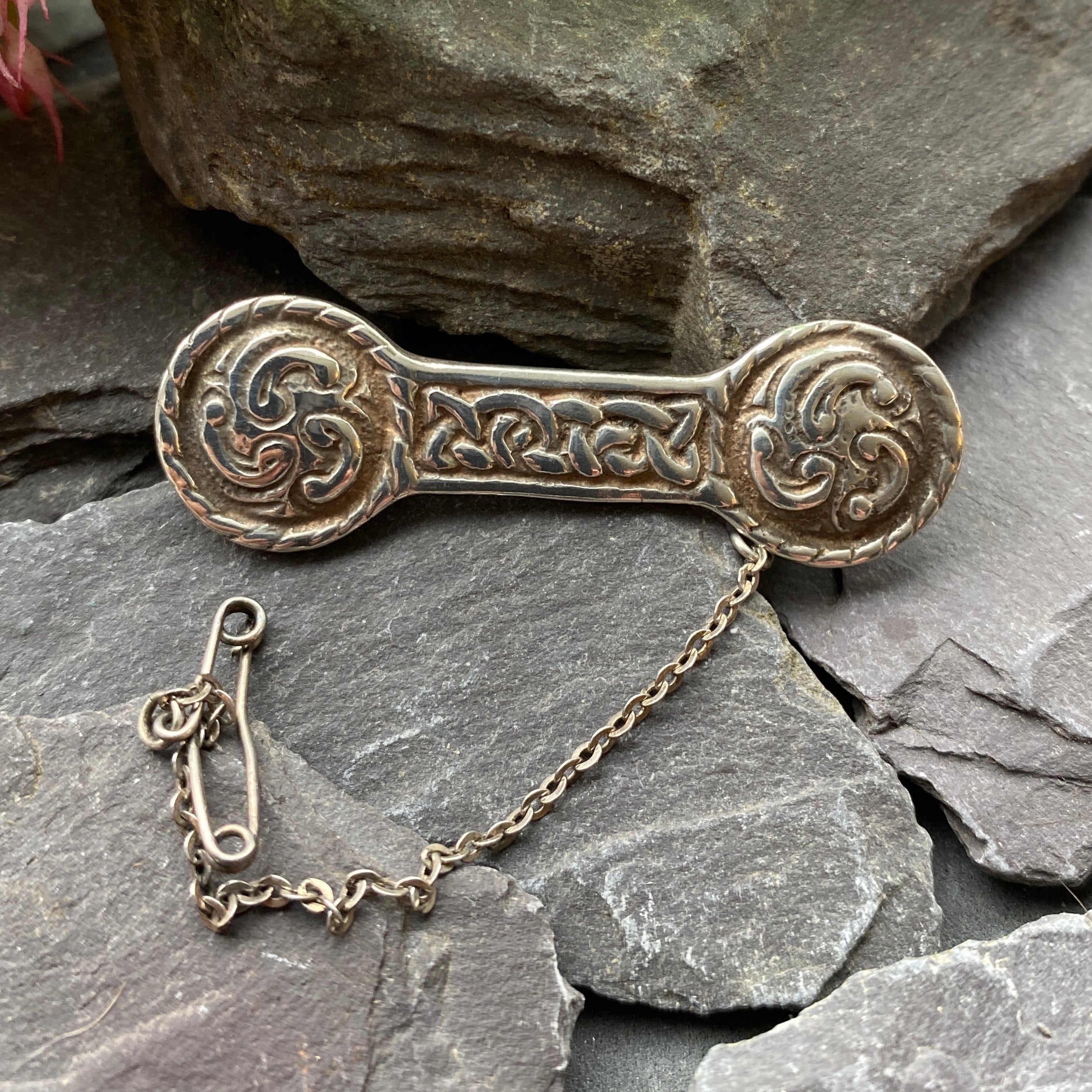Decorative silver brooch on a stone surface with green leaves and flowers.