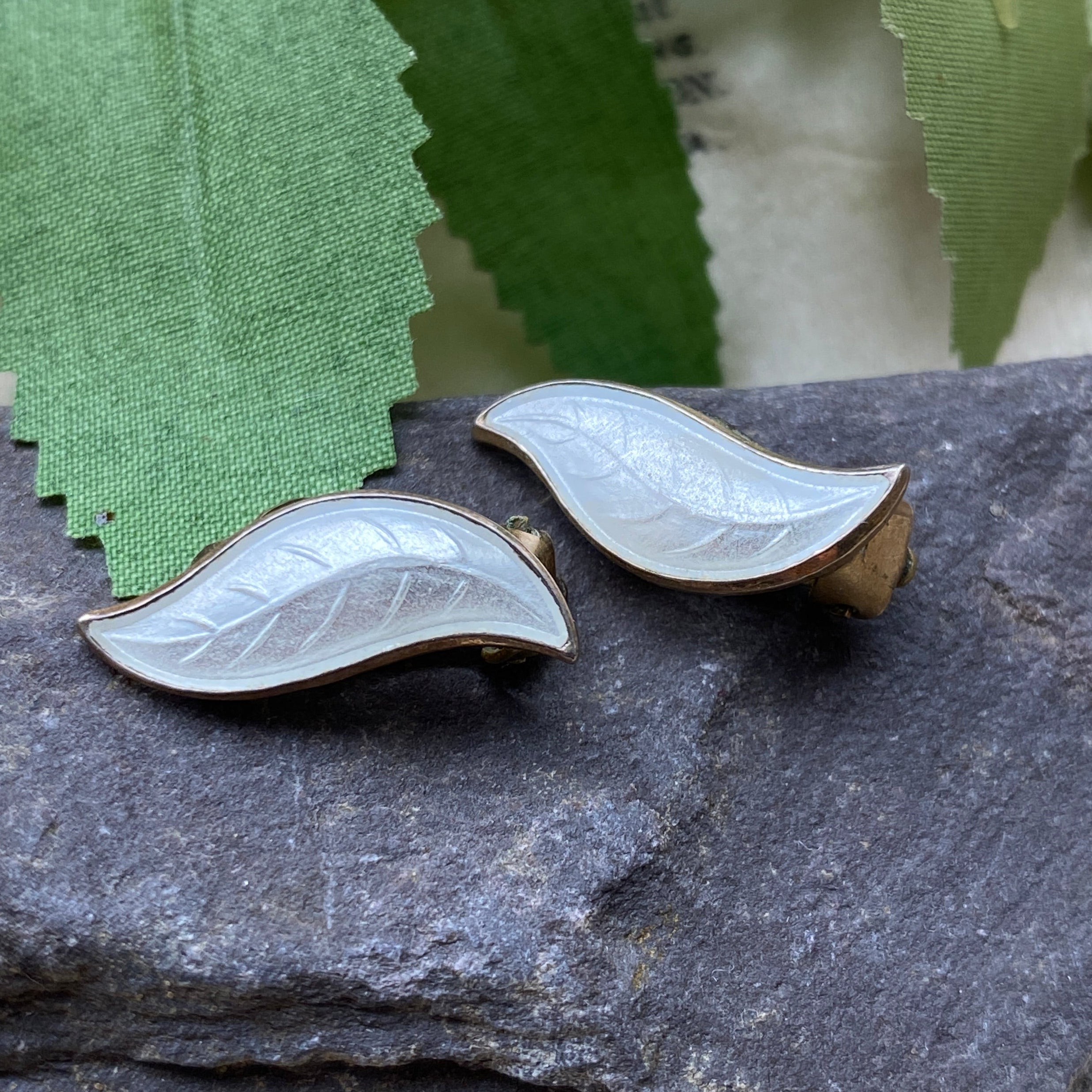Two leaf-shaped earrings on a dark stone surface with green leaves and pink flowers in the background.