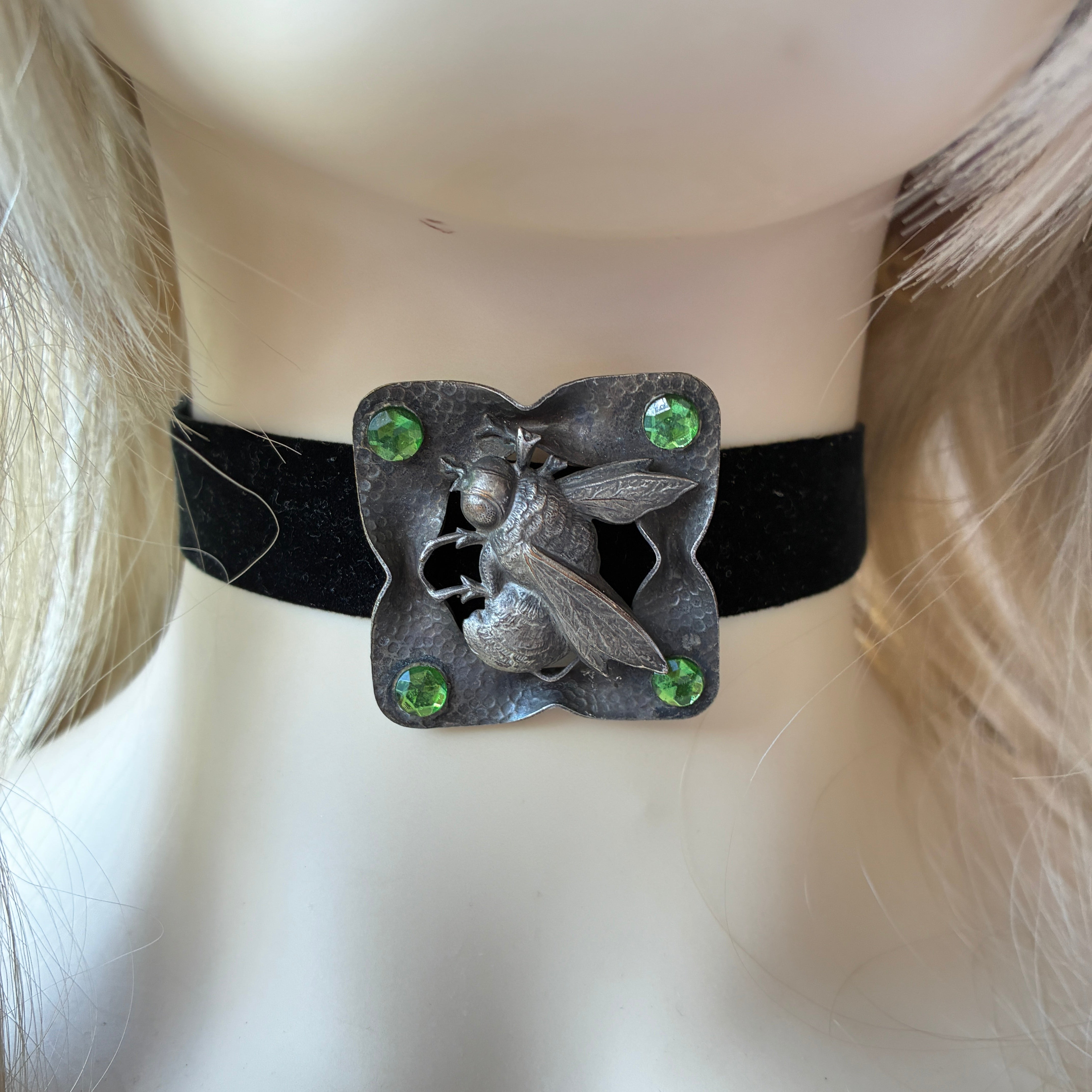A close-up of a black velvet choker necklace with a large, ornate Pewter Bee-shaped clasp with four green gemstone accents, displayed on a mannequin bust with blonde hair.